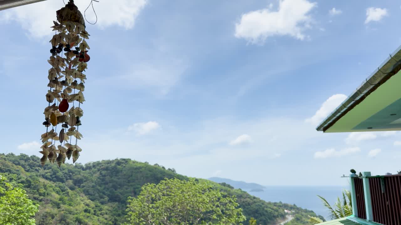 Un carillón de viento de conchas marinas se mece suavemente contra un exuberante paisaje montañoso con vistas al océano en el soleado Phuket, Tailandia