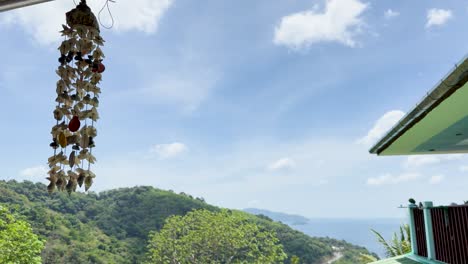 seashell wind chime sways gently against a lush, hilly backdrop with ocean views in sunny phuket, thailand
