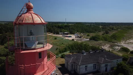 beautiful revealing aerial view of white painted steel lighthouse with red top located in pape, latvia at baltic sea coastline in sunny summer day, wide angle drone shot moving bacwards close