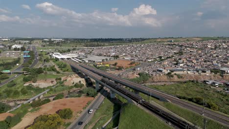 Clip-Aéreo-De-4k,-Un-Dron-Estático-Captura-La-Energía-Dinámica-De-Un-Tren-De-Alta-Velocidad,-Yuxtapuesto-A-Las-Bulliciosas-Carreteras-Y-Al-Extenso-Municipio-Urbano-De-Alexandra-En-Johannesburgo,-Sudáfrica