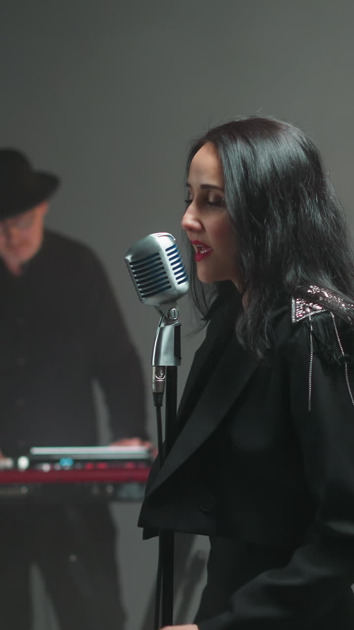 검은 옷을 입은 보컬이 빈티지 마이크에서 열정적으로 노래하고, 키보디스트는 배경에서 흐릿하게 보입니다.