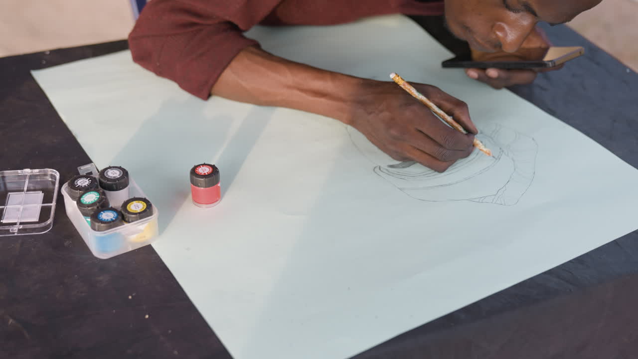 Hands drawing dotted design on paper, studio scene with pale sheet on black mat, small jars of pigment and ink, pencil tracing circular pattern, focused maker shaping mandalalike motif, quiet