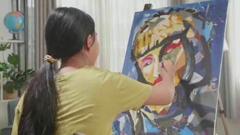 hind view of an asian artist girl in wheelchair holding paintbrush and painting a girl's face on the canvas