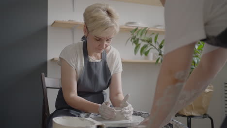 A-female-potter's-wheel-sculpting-teacher-explains-how-to-work-and-teaches-an-elderly-woman-to-work-with-clay-and-make-mugs-and-jugs.-Master-class-for-pensioners.-Pottery-courses