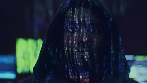 close up of the young guy hacker in a hood looking straight to the camera with projected code numbers and characters on his face in the dark room full of computers