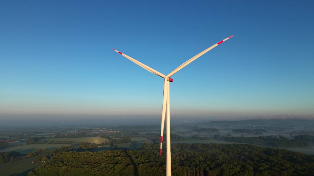 Una singola turbina eolica che ruota contro un cielo azzurro limpido durante l'alba dell'ora d'oro, con nebbia mattutina su un quartiere periferico e un paesaggio verde in un ambiente rurale americano. Orbita aerea ravvicinata.