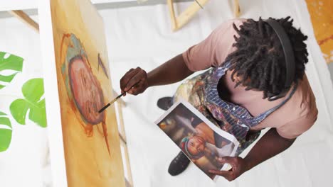 overhead view of african american male artist painting on canvas looking at photo at art studio