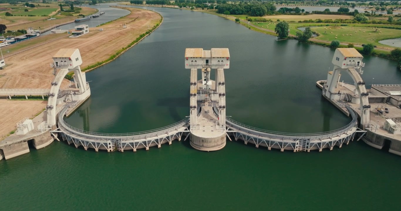 Drone approaches a hydroelectric power plant head-on, showcasing its distinctive arch dam, calm canal water and impressive renewable energy infrastructure on a sunny day.