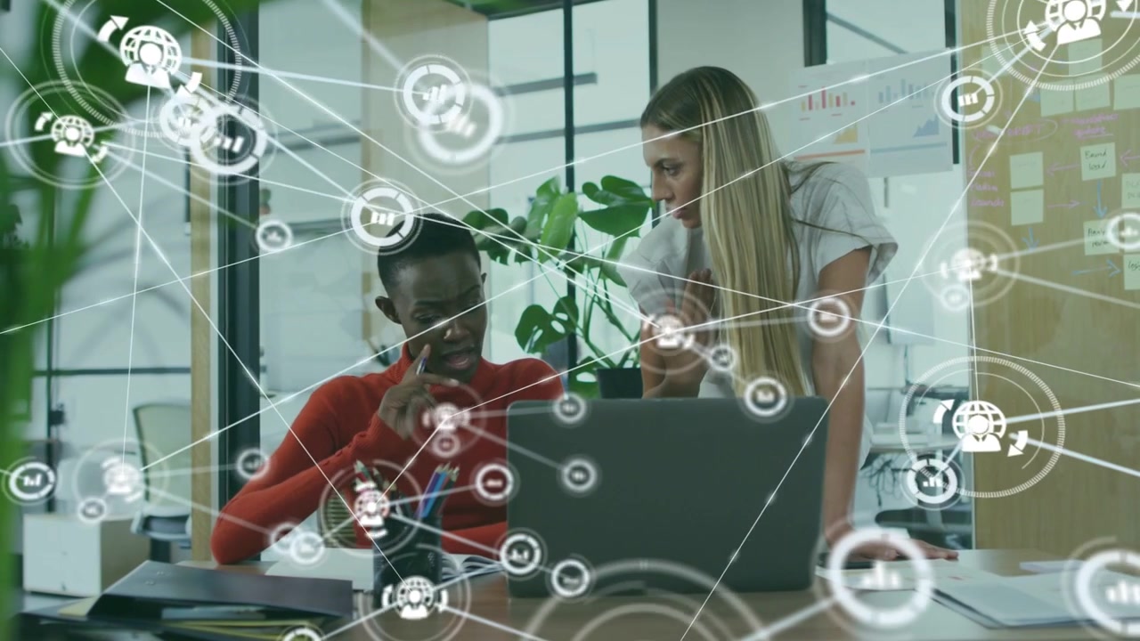 Diverse female coworkers leaning over laptop at desk red sweater pointing at screen with data map