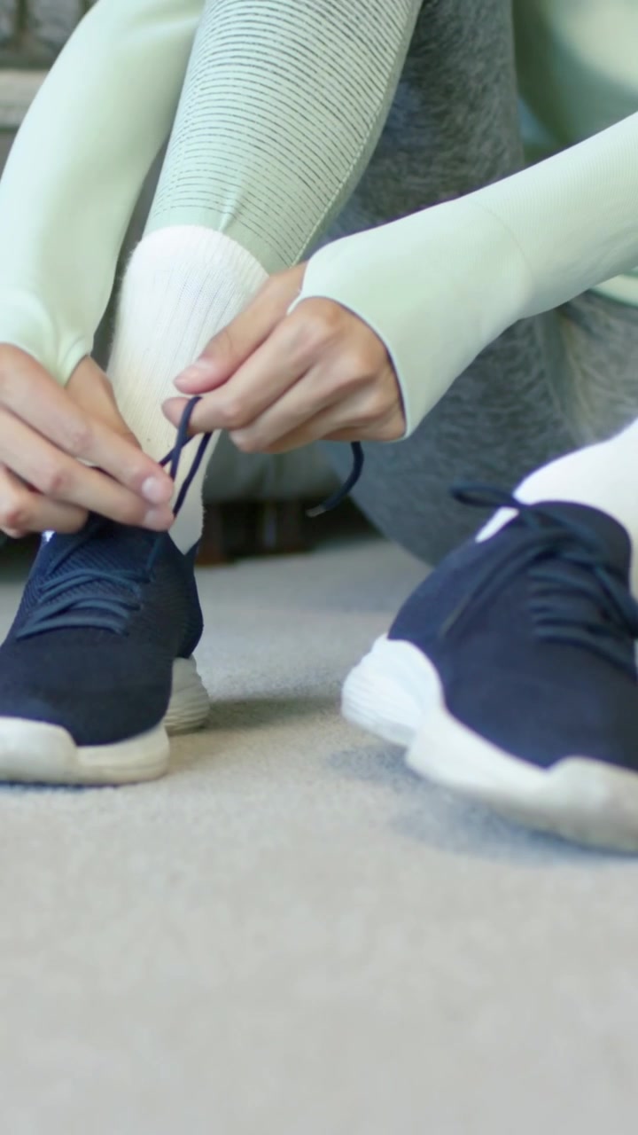 Video verticale: Una donna con top color menta e leggings che allaccia una scarpa blu navy sul tappeto di casa, preparandosi per l'allenamento.
