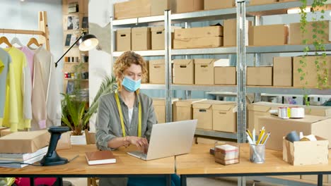 Young-caucasian-designer-woman-in-facial-mask-sitting-at-desk-and-videochatting-on-laptop-in-clothing-shop-warehouse