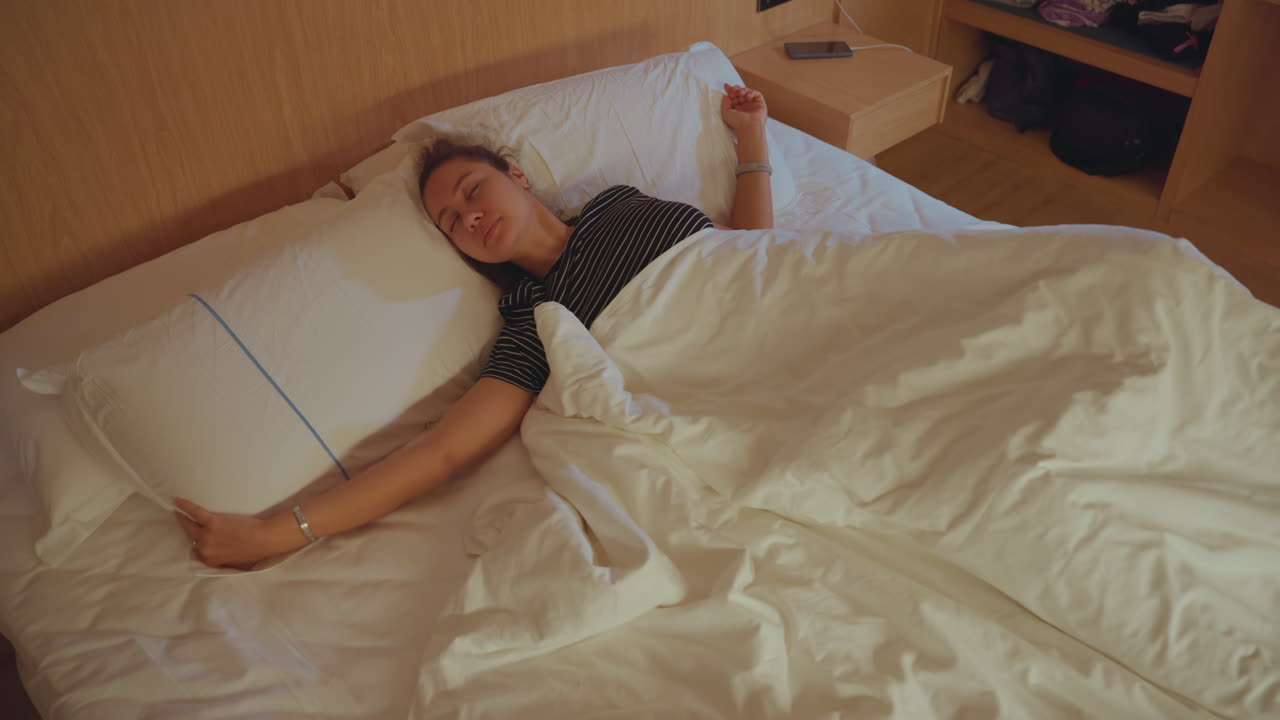 Kvinde slapper af alene. Kvinden begynder dagen i et fredfyldt hotelmiljø. Enlig kvindelig gæst strækker sig på sengen med blid belysning. Individuel kvinde på hotellet finder ro i de fredelige omgivelser