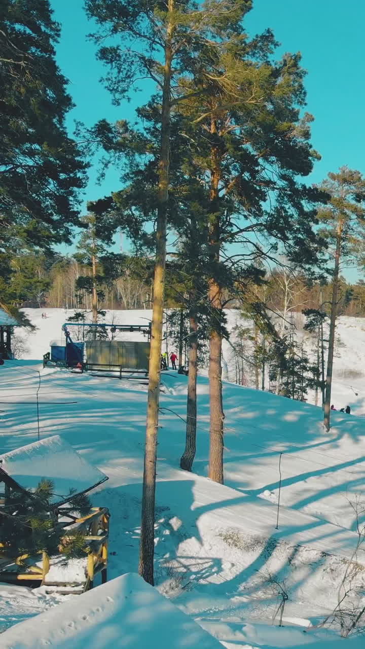 wooden gazebo covered with snow against people on mountain ski resort behind green pine trees on sunny day aerial view