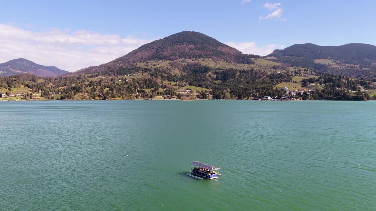 Vista aérea de drone de um barco navegando por águas cintilantes com montanhas em camadas ao fundo, no Lago Colibita, Romênia