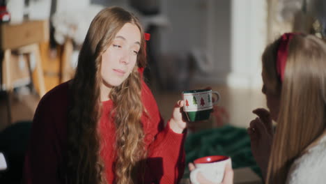 Pretty-woman-with-coffee-cup-talking-to-sister-at-bright-home-during-Christmas