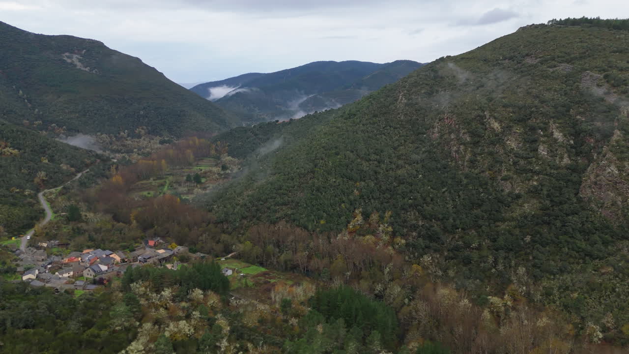 Aerial view of a small mountain village surrounded by dense green hills and autumn trees, with morning mist drifting through the valley under cloudy skies