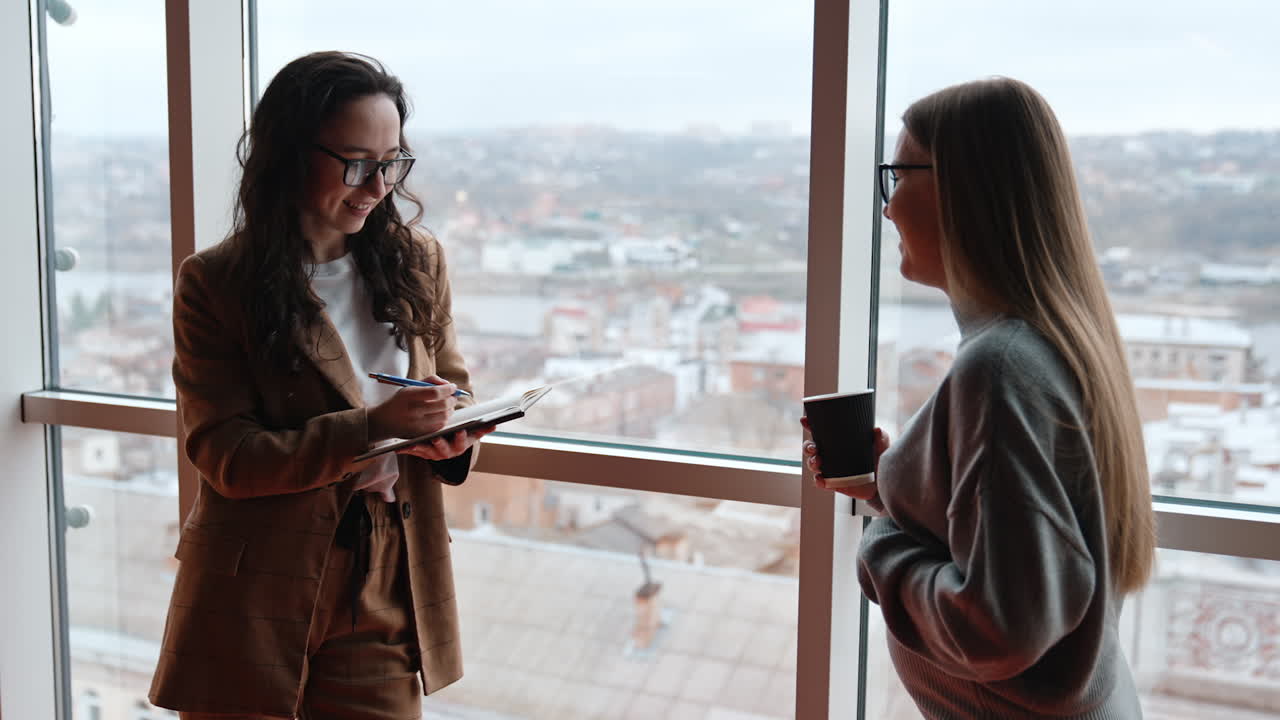 Young ladies talk in the office standing near the panoramic window. Brunette has a notebook in hand and blonde one drinking coffee.