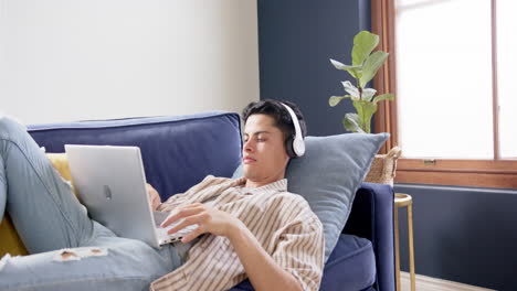 focused biracial man wearing headphones lying on couch using laptop at home, copy space, slow motion