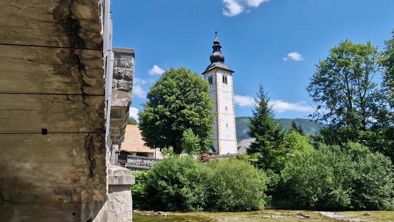 Katolsk kirke ved Bohinj-sjøen, Slovenia. Tårn og bro over elveutløp på en varm solskinnsdag