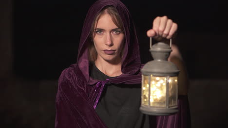 mysterious role play woman wearing a purple cape and hood and holding a lantern at night