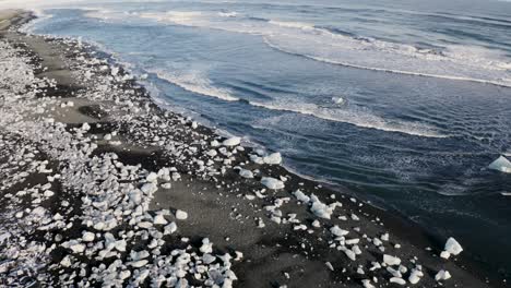 aerial shot of crystal lagoon’s dramatic coastline in iceland, where glacial ice fragments scatter across black volcanic sand, contrasting with cold blue waves in motion.