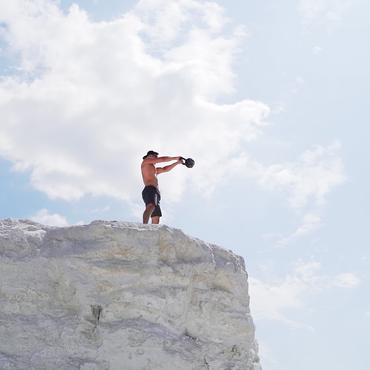 Strong man lifting kettlebell on the mountain in summer. Male athlete swinging kettlebell with two hands on the beautiful natural background outdoors.