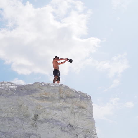 strong man lifting kettlebell on the mountain in summer. male athlete swinging kettlebell with two hands on the beautiful natural background outdoors.
