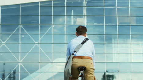 Back-view-of-attractive-man-in-the-casual-style-riding-a-bicycle-close-to-a-glass-building