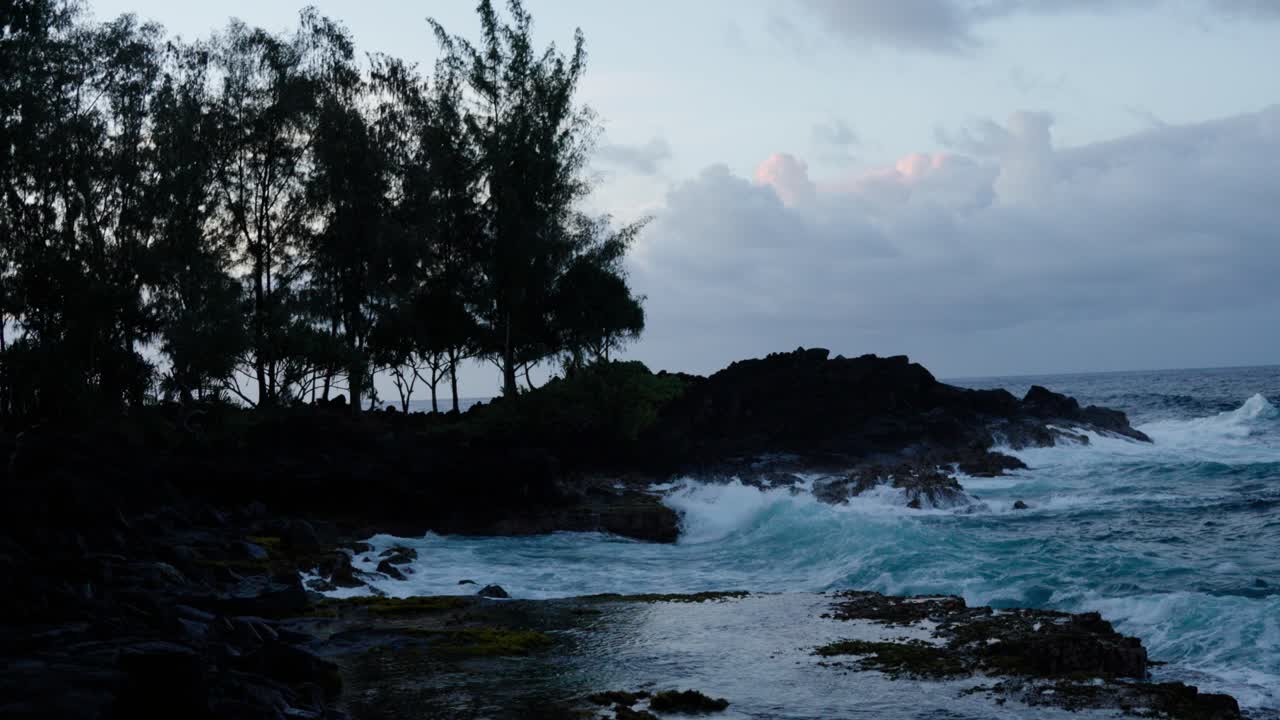 Powerful ocean waves break against dark volcanic rocks at dusk, silhouetted trees lining the shoreline under soft twilight clouds.