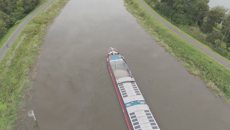 Drone-shot-of-a-Barge-sailing-over-a-Belgian-river-during-daytime-with-grey-weather-with-trees-and-nature-in-the-background-and-a-small-city-LOG