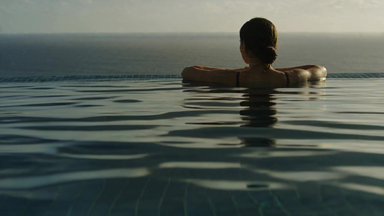 Blickende Frau mit dunklen Trägern entspannt am Rand eines Infinity-Pools, Meer, Sonne und Brise bewegen Wellen.