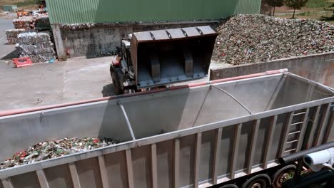 forklift excavator is sipping glass into back of an transporter truck in warehouse, heavy-duty machine has a long arm fitted with a glass-grabbing attachment, drone view, glass recycling concept