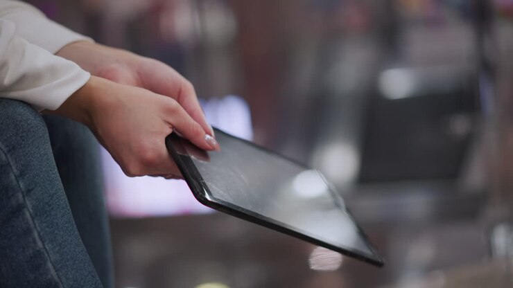 Close up of hands with polished nails gently swaying tablet, showcasing subtle light reflections on screen and blurred colorful background
