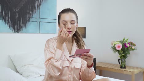cute woman applying eye shadows in pink pajamas in the white bedroom