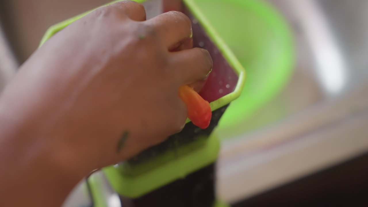 Closeup of hand filling green handcrank grinder with cherry tomatoes over sink, gentle pressing and turning motion, water droplets on metal sink, home cook preparing fresh tomato sauce and seasoning