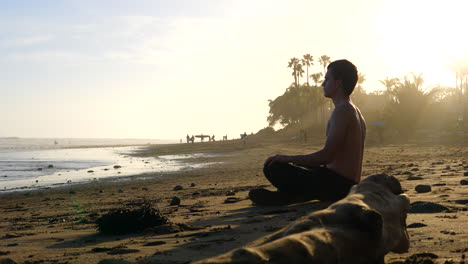 a handsome man meditating and watching surfers at sunset on a picturesque california beach with palm trees and people in silhouette