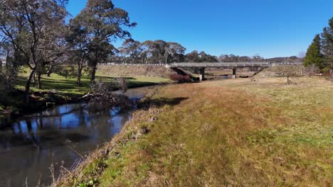 drone glides above a winding stream, grassy banks, and a rustic bridge near ebor falls, new south wales, under clear daylight with steady camera movement