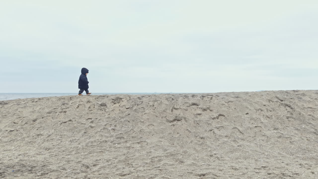 Child walks alone on the beach at a cloudy day with gray skies and soft sand near the water's edge