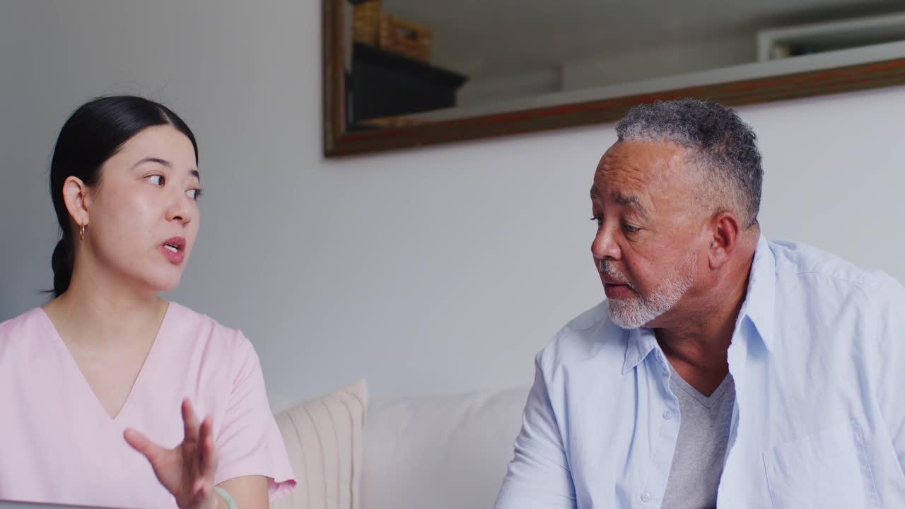 Asian caregiver in pink scrub reading tablet and explaining care to African American man on sofa