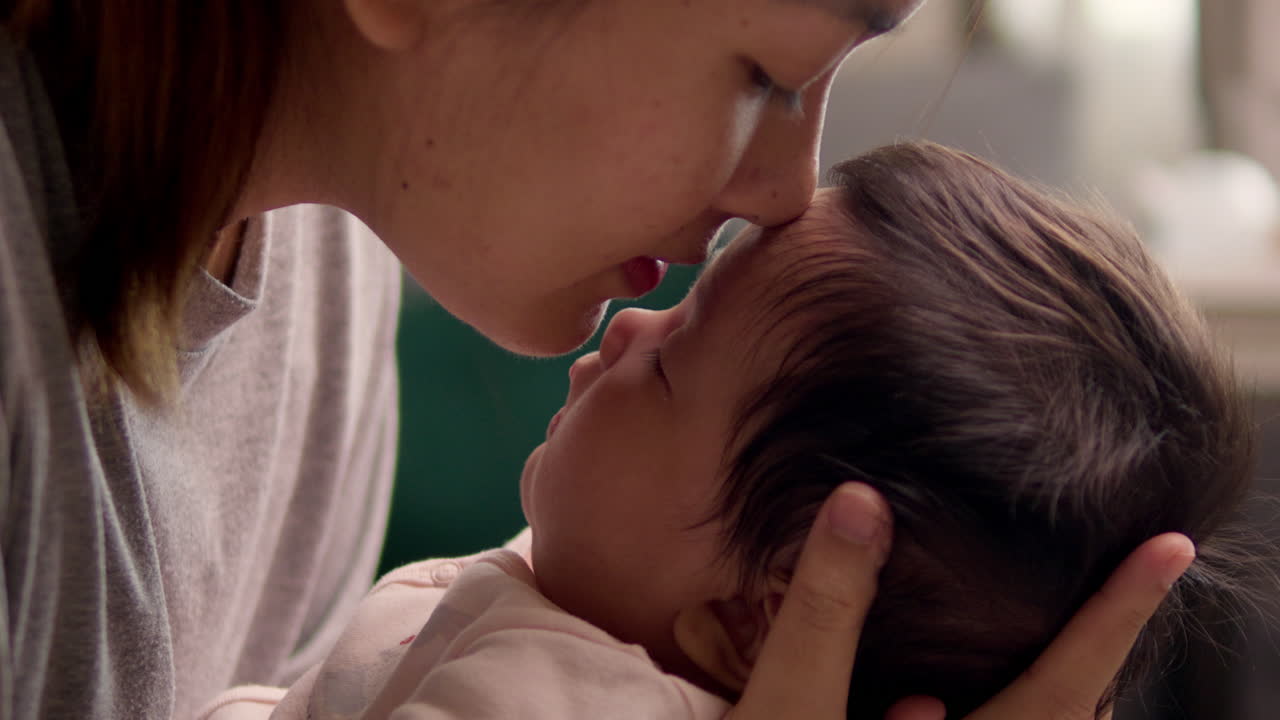 A mother leans down to smell her newborn child's head and gently kisses her baby on the forehead.