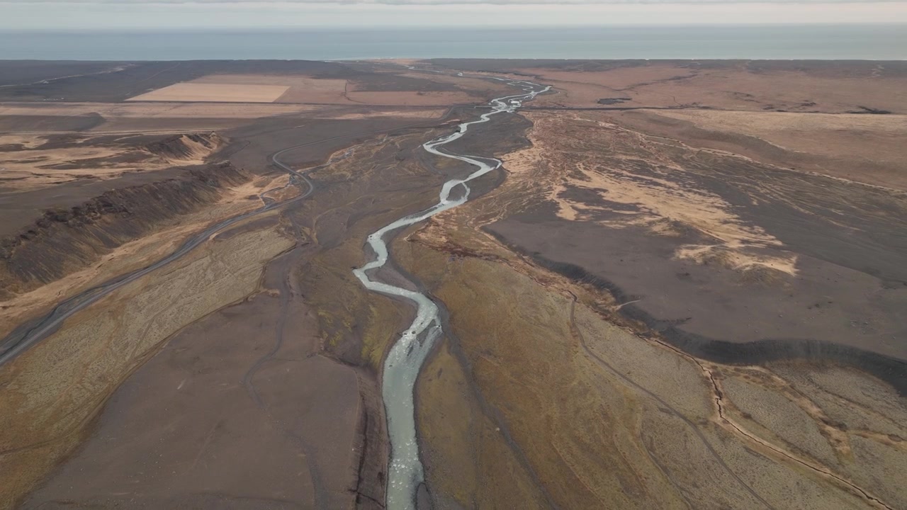 Ripresa con drone di un fiume serpeggiante che attraversa le pianure desolate dell'Islanda meridionale, con terreno accidentato e campi agricoli distanti sullo sfondo