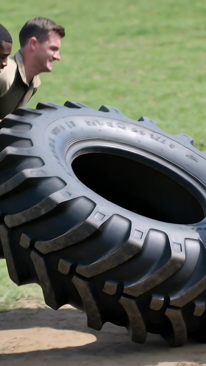 Vertical video: Surging from crouch two soldiers pushing tire across training field in camo gear