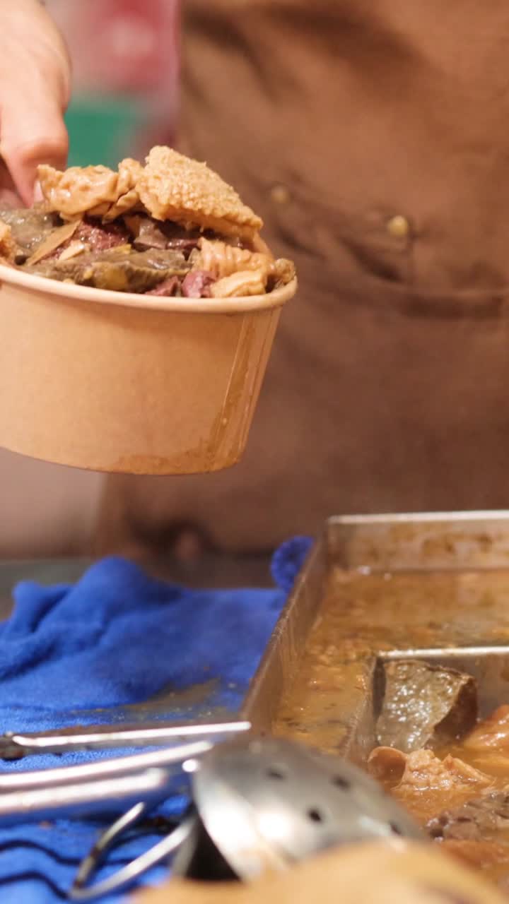 Um vendedor de comida de rua usa pinças para encher uma tigela de papel com miúdos cozidos saborosos.