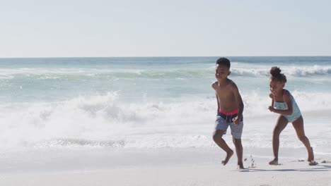 hermanos afroamericanos sonrientes corriendo y usando trajes de baño en una playa soleada