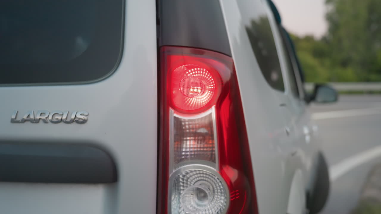 Close up of motor car making turn with taillight illuminated as another car passes in blurred background along roadside, silver vehicle reflecting evening light with calm greenery surroundings