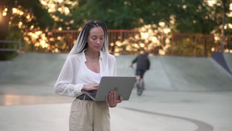 mujer joven elegante con rastas de pelo blanco y negro caminando por el parque o skatepark con portátil plateado abierto, escribiendo.