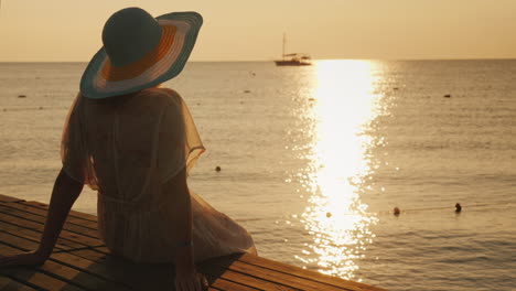 Una-Mujer-Joven-Se-Encuentra-Con-El-Amanecer-En-El-Muelle,-Se-Sienta-Y-Mira-El-Sol-Y-El-Barco-En-El-Mar-Dre