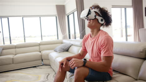 un joven biracial disfruta de un juego de realidad virtual con espacio de copia, sentado en un sofá en casa