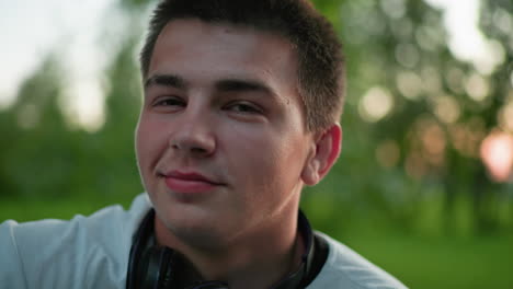 primer plano de un hombre con auriculares negros alrededor del cuello, con una camiseta blanca y sentado al aire libre con una cálida sonrisa, rodeado de árboles verdes y una suave iluminación al aire libre