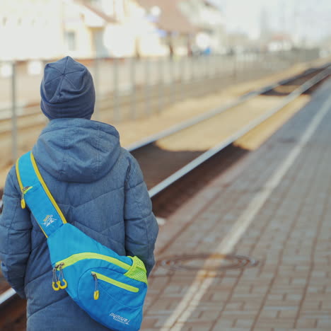 young boy in winter clothes waiting for the train on the background of a railway station. back view of one boy with a bag on his back standing on the platform and looking ahead.